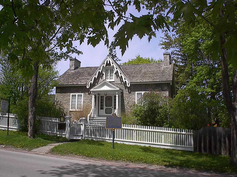 A short, grey brick house with a white picket fence surrounded by trees and a green lawn. 